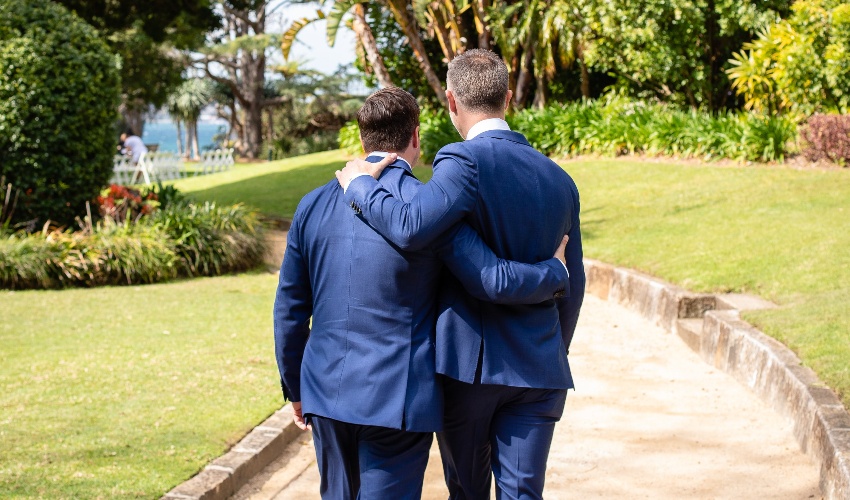 Two men in blue suits walking down a walkway during a tropical wedding