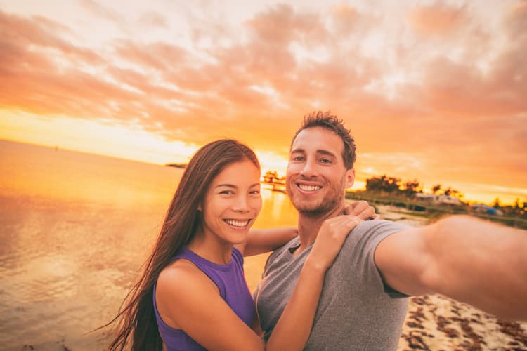 Happy selfie couple tourists on USA travel taking photo at sunset on Florida beach.