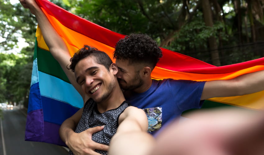 two men embrace and hold up a rainbow flag at a Pride parade