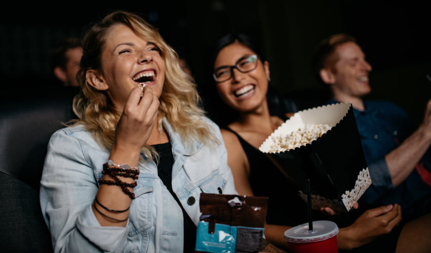 two women laugh and eat popcorn as they watch a film in a movie theater