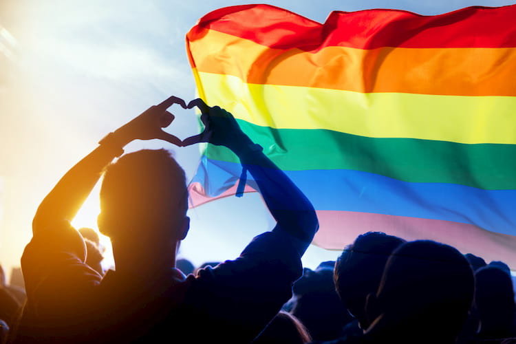 someone makes a heart shape with their hands in front of a rainbow pride flag