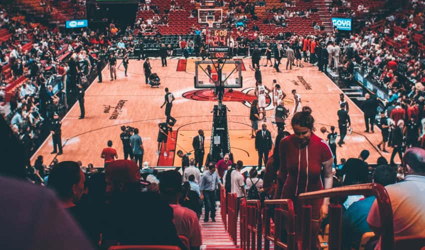 Fans, coaches, and players walk around a basketball court before a game, the Miami Heat logo visible in the middle of the court