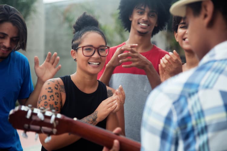 Young people listening and clapping while man plays guitar