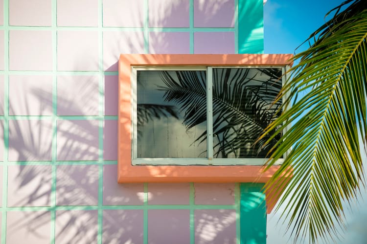a corner window with an orange trim against a pink tile building with palm leaves in the foreground