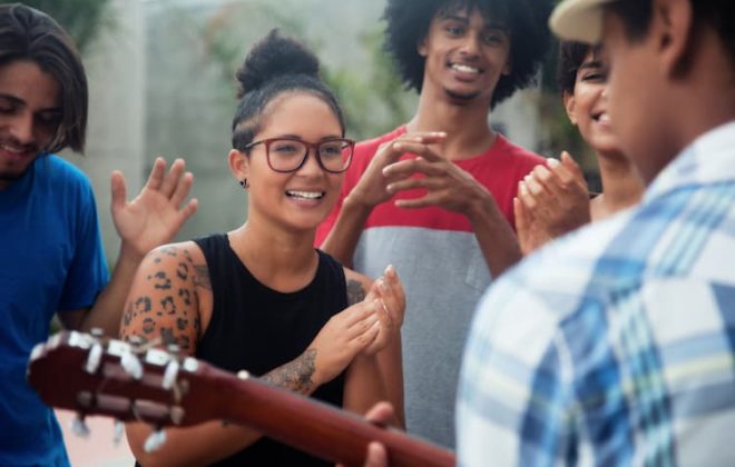 Young people listening and clapping while man plays guitar